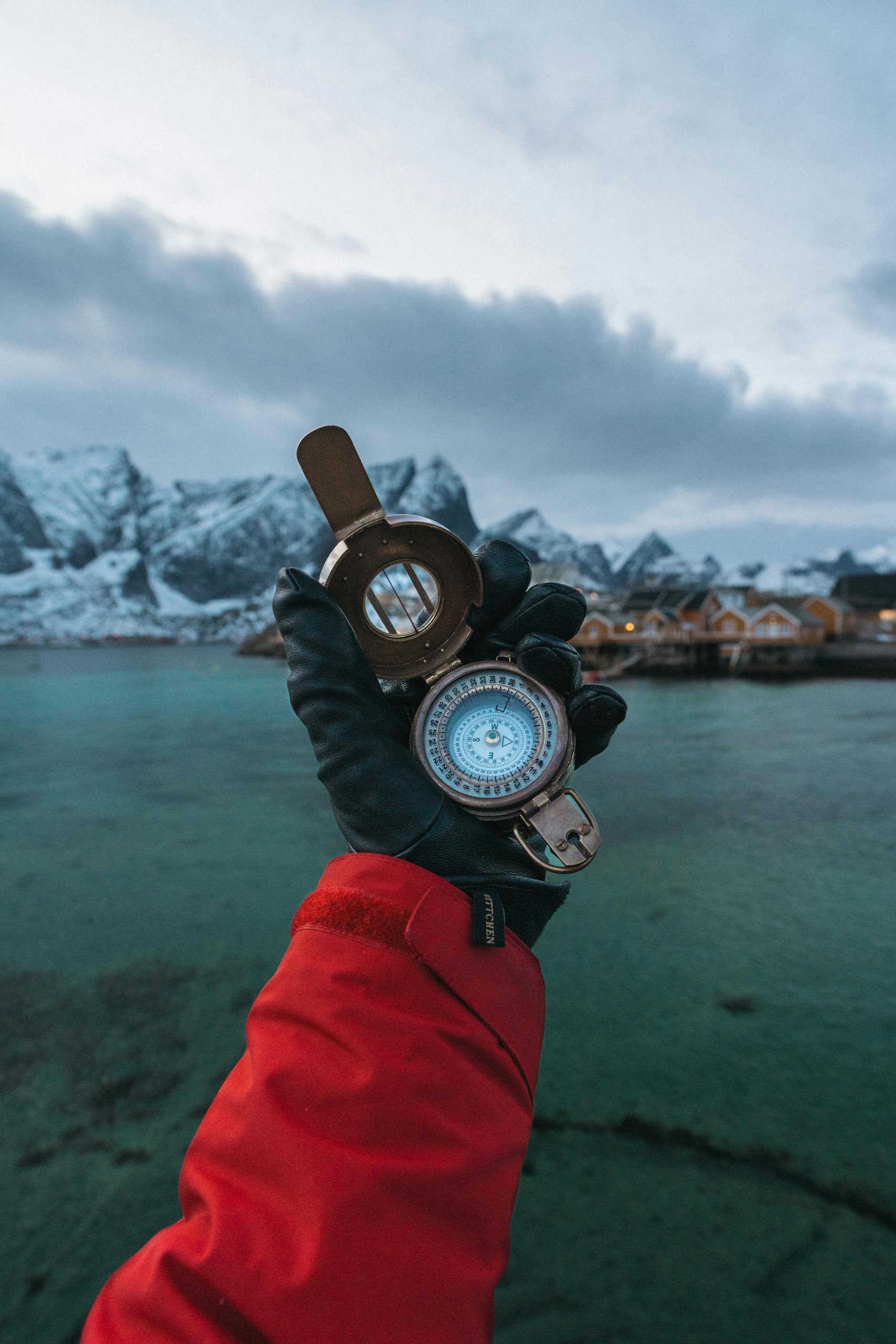 A gloved hand holds a compass in Reine, Lofoten Islands, with a stunning fjord backdrop.