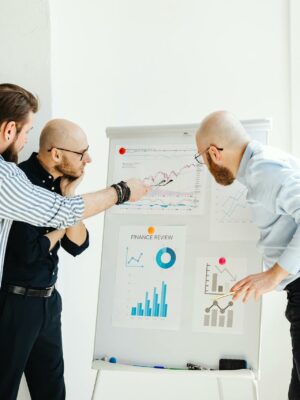 Photo by Karola G Three colleagues in a bright office discuss financial charts on a whiteboard.